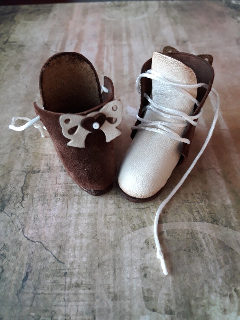May include: A pair of miniature leather boots with laces. One boot is brown suede with a white bow detail, the other is white leather.