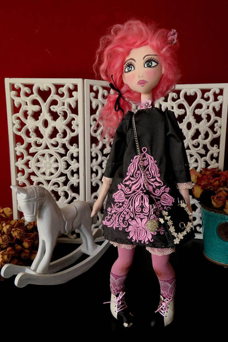 May include: A handmade doll with pink hair, wearing a black dress with pink floral embroidery, black shoes, and pink tights. The doll is holding a black purse with a gold chain and white flowers.