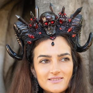 May include: A woman wearing a black horned headdress with a skull and red accents. She is also wearing a black choker and a black corset. The headdress features black horns, crystals, and a skull with red eyes.