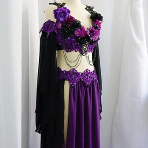 May include: A purple and black belly dance costume on a mannequin. The top is black with purple floral accents and beaded detailing. The skirt is a flowing purple fabric. Black sleeves are attached to the top.