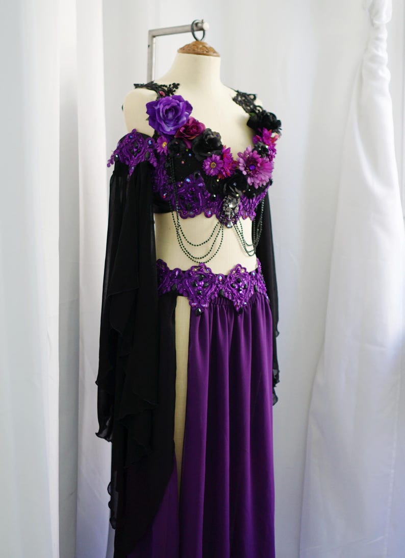 May include: A purple and black belly dance costume on a mannequin. The top is black with purple floral accents and beaded detailing. The skirt is a flowing purple fabric. Black sleeves are attached to the top.