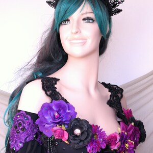 May include: A black lace crown with purple and blue accents sits atop a mannequin head with teal hair. The mannequin wears a black and purple costume adorned with large purple and black flowers and beaded embellishments.