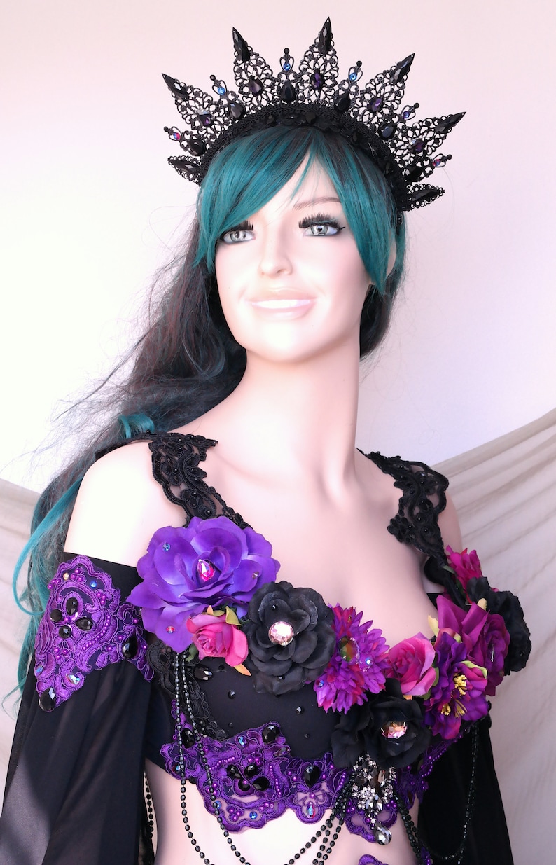 May include: A black lace crown with purple and blue accents sits atop a mannequin head with teal hair. The mannequin wears a black and purple costume adorned with large purple and black flowers and beaded embellishments.
