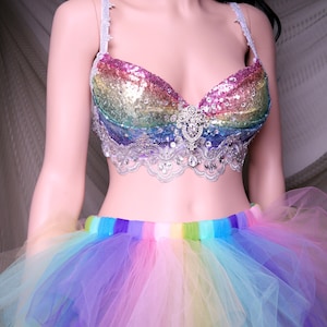 May include: A rainbow sequined bra with a white lace trim and a large rhinestone embellishment in the center. The bra is paired with a rainbow tulle skirt.