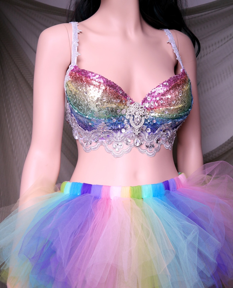 May include: A rainbow sequined bra with a white lace trim and a large rhinestone embellishment in the center. The bra is paired with a rainbow tulle skirt.
