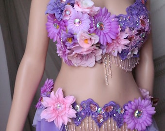 Puede incluir: Un disfraz de danza del vientre de dos piezas con un diseño floral. La parte superior está adornada con flores moradas, rosas y blancas, adornadas con pedrería y cuentas colgantes. El cinturón a juego tiene un diseño floral y de pedrería similar, con una falda morada fluida.