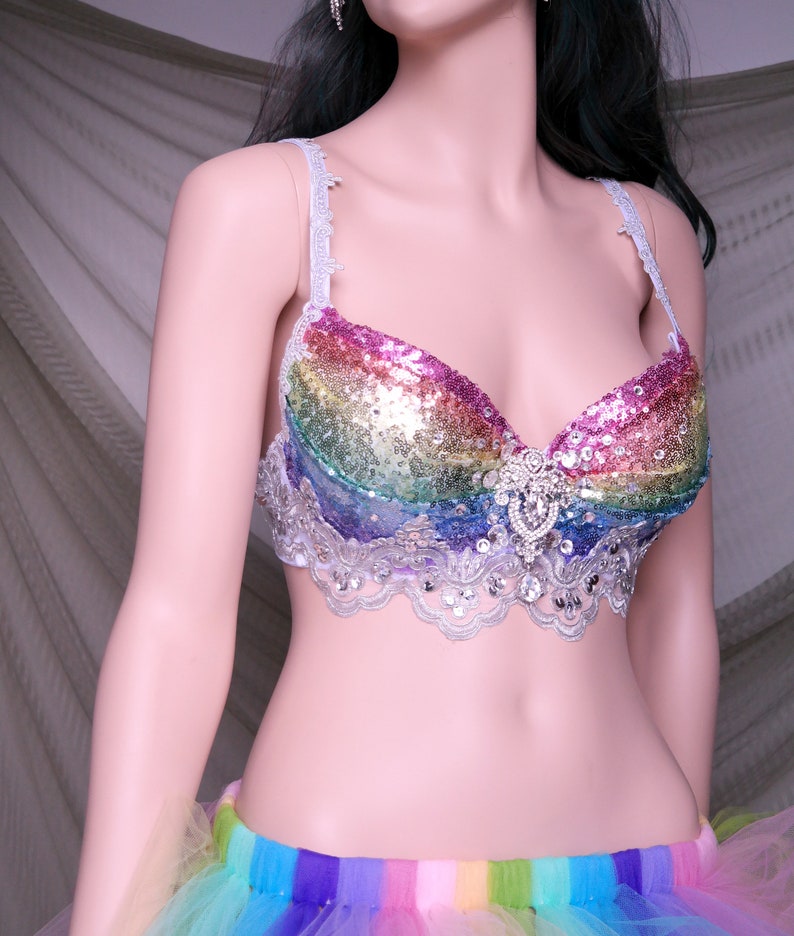May include: A rainbow sequined bra with a silver lace trim and a large rhinestone embellishment in the center. The bra is worn over a rainbow tulle skirt.