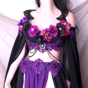 May include: A black and purple belly dance costume with floral accents. The top is adorned with purple and black flowers, and the skirt features a high slit. The costume includes beaded embellishments and flowing black sleeves.