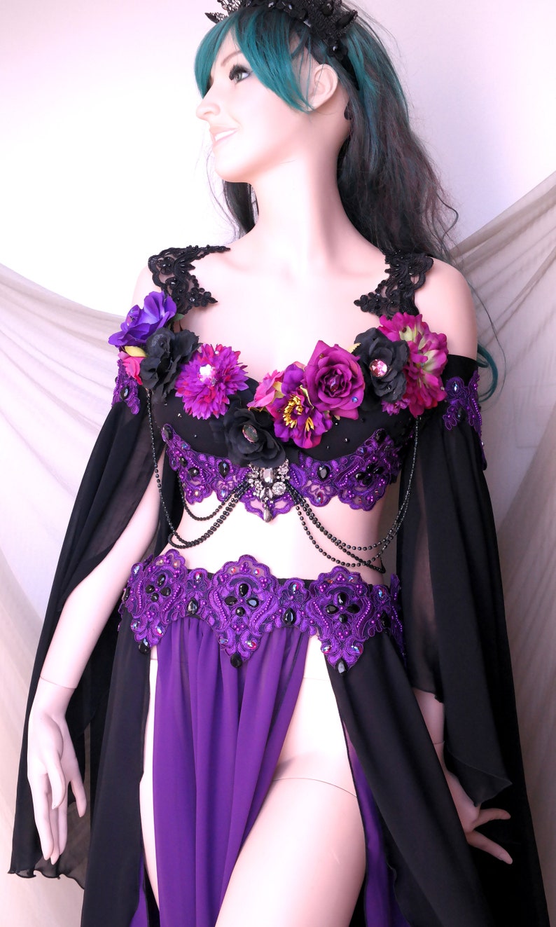 May include: A black and purple belly dance costume with floral accents. The top is adorned with purple and black flowers, and the skirt features a high slit. The costume includes beaded embellishments and flowing black sleeves.