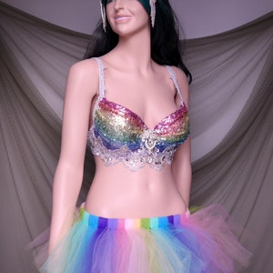 May include: A rainbow sequined bra with a white lace trim and a rainbow tulle skirt. The bra has a silver rhinestone embellishment in the center. The skirt has a rainbow striped waistband.