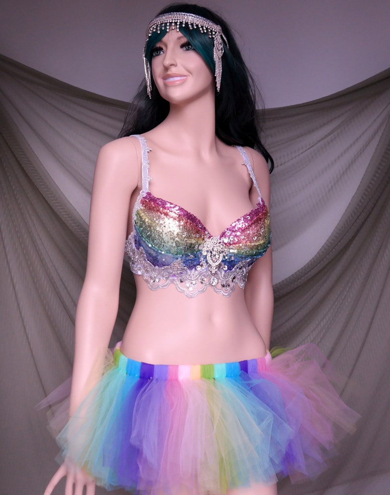 May include: A rainbow sequined bra with a white lace trim and a rainbow tulle skirt. The bra has a silver rhinestone embellishment in the center. The skirt has a rainbow striped waistband.