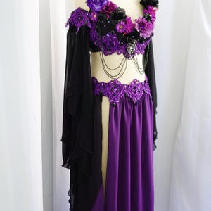 May include: A purple and black belly dance costume on a mannequin. The top is embellished with purple and black flowers and beads. The skirt is a long, flowing purple with a slit. Black sleeves drape from the shoulders.