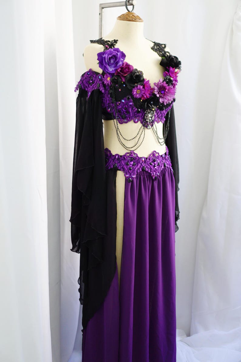 May include: A purple and black belly dance costume on a mannequin. The top is embellished with purple and black flowers and beads. The skirt is a long, flowing purple with a slit. Black sleeves drape from the shoulders.