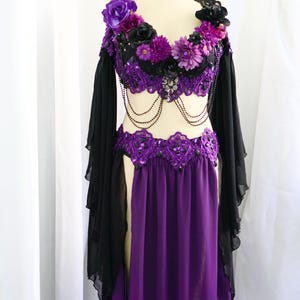 May include: A purple and black belly dance costume on a mannequin. The top is black with purple floral accents and beaded detailing. The skirt is a flowing purple fabric. Black sheer sleeves are attached to the top.