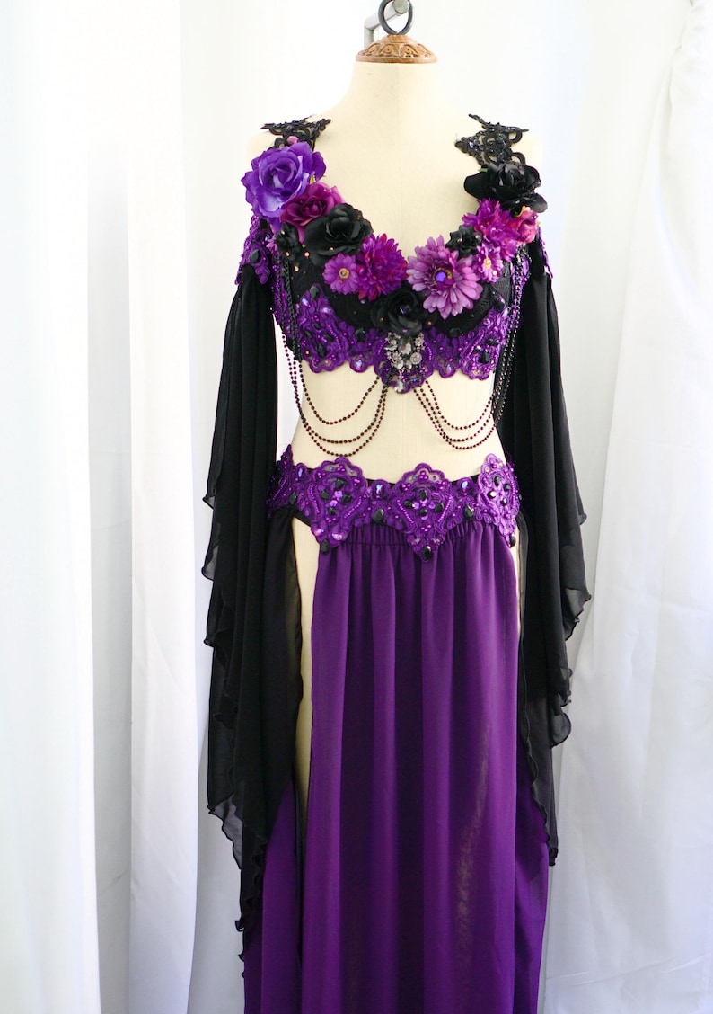 May include: A purple and black belly dance costume on a mannequin. The top is black with purple floral accents and beaded detailing. The skirt is a flowing purple fabric. Black sheer sleeves are attached to the top.