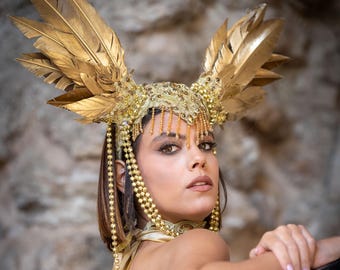 May include: A woman wearing a gold feathered headdress with beaded strands and a gold necklace. She is looking over her shoulder with a serious expression.