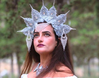 May include: A silver-colored, ornate headpiece with multiple points and dangling crystal accents. The headpiece is worn with a matching necklace and earrings. The woman is wearing a white dress, and the background is blurred green foliage.