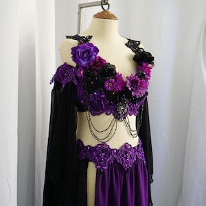 May include: A purple and black belly dance costume on a mannequin. The top is adorned with purple and black flowers, lace, and beaded accents. The skirt is a deep purple color, and the costume has long black sleeves.