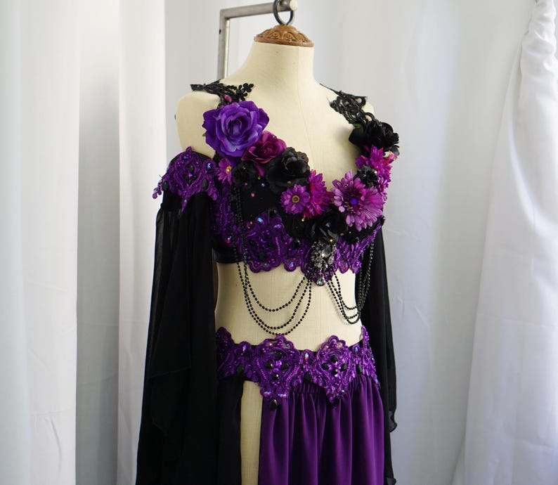 May include: A purple and black belly dance costume on a mannequin. The top is adorned with purple and black flowers, lace, and beaded accents. The skirt is a deep purple color, and the costume has long black sleeves.