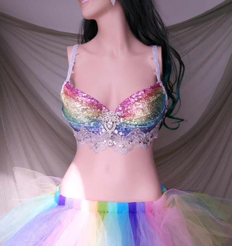 May include: A rainbow sequined bra with a white lace trim and a large rhinestone embellishment in the center. The bra is paired with a rainbow tulle skirt.