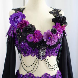 May include: A black and purple belly dance costume with floral and beaded embellishments. The top features a black base with purple lace trim, adorned with purple and black flowers. Long black sleeves and beaded accents complete the look.