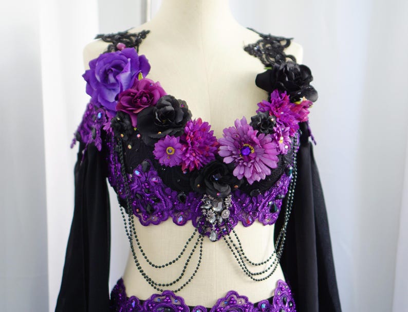 May include: A black and purple belly dance costume with floral and beaded embellishments. The top features a black base with purple lace trim, adorned with purple and black flowers. Long black sleeves and beaded accents complete the look.