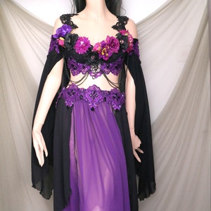 May include: A black and purple costume with floral accents. The off-the-shoulder top is adorned with purple and pink flowers and black lace. The costume includes a black cape and a long, sheer purple skirt. A black crown with spikes completes the look.
