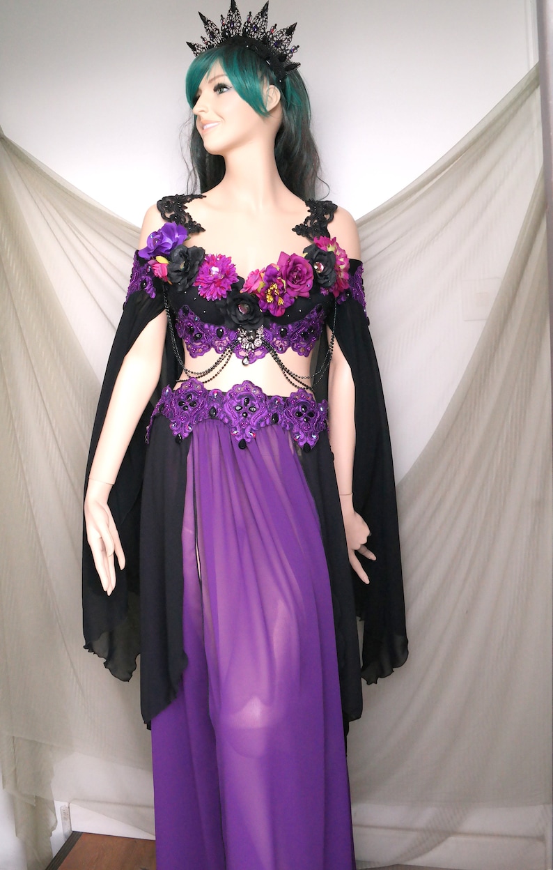 May include: A black and purple costume with floral accents. The off-the-shoulder top is adorned with purple and pink flowers and black lace. The costume includes a black cape and a long, sheer purple skirt. A black crown with spikes completes the look.