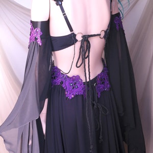 May include: Black belly dance costume with purple and black embellishments. The costume features a black bra, a skirt, and sheer sleeves. The back is open with a rope tie. The purple accents include beaded designs and lace.
