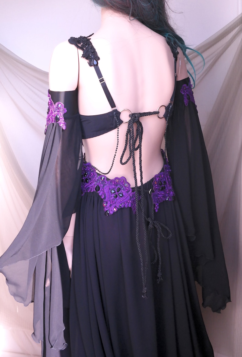 May include: Black belly dance costume with purple and black embellishments. The costume features a black bra, a skirt, and sheer sleeves. The back is open with a rope tie. The purple accents include beaded designs and lace.