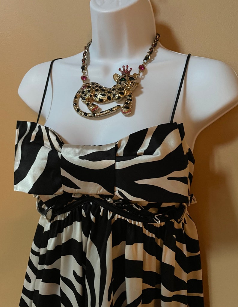 May include: A black and white zebra print dress with a black satin ribbon around the waist. The dress has a sweetheart neckline and thin straps. A gold necklace with a jeweled leopard pendant is worn on a mannequin.