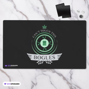 May include: Black rectangular playmat with the text "CAN'T TOUCH THIS" and "BOGLES" in white and green. The playmat has a circular design with arrows and a crown. Two black card sleeves and dice are in the background.