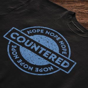 May include: Black t-shirt with a blue distressed circle stamp that says "NOPE NOPE NOPE" around the word "COUNTERED" in the center.