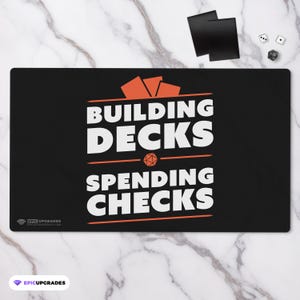 May include: Black rectangular gaming mat with the text "BUILDING DECKS SPENDING CHECKS" in white and orange. The mat has a logo in the bottom left corner and is on a marble surface. Dice and card sleeves are in the background.