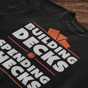 May include: Black t-shirt with white text that reads "Building Decks Spending Checks". The text is in a bold, sans-serif font. There is a stylized orange dice graphic in the middle of the text.