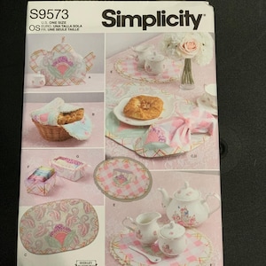 May include: A Simplicity S9573 sewing pattern envelope featuring instructions for fabric kitchen accessories. The cover displays a tea cozy, bread basket cover, napkins, placemats, and small fabric baskets in pink, white, and green floral and gingham patterns.