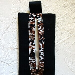 May include: A rectangular black fabric tissue holder featuring a central vertical opening trimmed with a brown, black, and white animal print pattern. The holder includes a small black fabric loop at the top for hanging or attachment purposes.