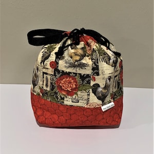 May include: A fabric drawstring bag featuring a print of roosters, flowers, and script text on a cream background. The bottom section is a solid red fabric with a black honeycomb pattern. It has a black nylon strap and black drawstring cords.