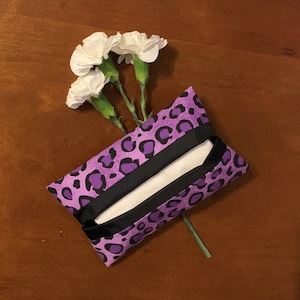 May include: A rectangular fabric tissue holder featuring a vibrant purple and black leopard print pattern with a black fabric center opening. Three white carnation flowers with green stems are tucked behind the tissue holder.