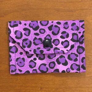 May include: A rectangular fabric pouch featuring a vibrant purple and black leopard print pattern. The pouch has a triangular flap closure secured with a single centered black plastic snap button. The edges are finished with visible black stitching.