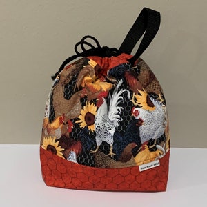 May include: A fabric drawstring bag featuring a pattern of chickens, roosters, and sunflowers against a black chicken wire background. The base is a solid red fabric with a hexagonal pattern. It has a black strap and a small white tag reading Sew Knot Idle.