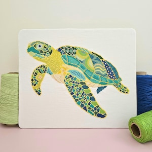 May include: A colorful needlepoint of a sea turtle, featuring shades of yellow, green, and blue. The turtle is set against a white background, with spools of green and blue thread on either side. The artwork is square and appears to be a finished piece.
