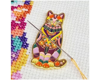 May include: A colourful enamel pin of a cat with geometric patterns in shades of purple, orange, yellow, and green. The pin is sitting on a white fabric background with a needle and thread.
