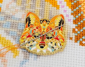 May include: A colourful fox pin with geometric patterns in shades of orange, red, yellow, blue, and white. The pin is made of metal and has a gold finish. A needle is threaded through the pin, suggesting it is a needle minder.
