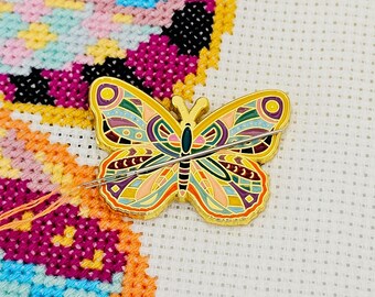May include: A gold butterfly-shaped needle minder with colorful geometric patterns and a needle threaded through it. The needle minder is sitting on a white Aida cloth.
