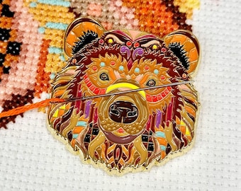 May include: A gold-colored metal bear pin with a colorful geometric design. The bear has a brown, orange, yellow, red, and purple design. The bear's eyes are black and the nose is brown.