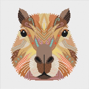 Op de afbeelding: Een kruissteekpatroon van een capybarakop met een symmetrisch ontwerp van geometrische vormen en lijnen in tinten bruin, beige, perzik en taling. Het dier heeft grote zwarte ogen en een donkerbruine neus, alles weergegeven op een wit raster.