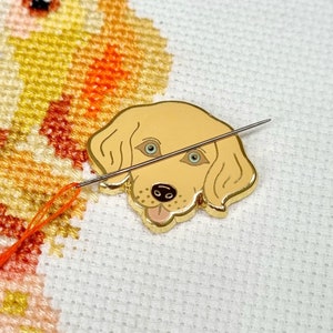 Puede incluir: Un pin de metal dorado de un perro de dibujos animados con ojos marrones y una lengua rosa. El pin está sobre un fondo de tela blanca con una aguja e hilo naranja.