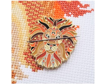 May include: A lion-shaped needle minder in shades of brown, orange, and gold, with a needle passing through it. The lion design features geometric patterns. The needle minder is on white fabric with cross-stitch.