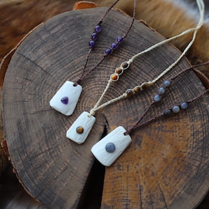 Puede incluir: Tres collares hechos a mano con colgantes rectangulares y detalles de cuentas. Cada colgante blanco tiene una incrustación de piedra preciosa. Los collares presentan cuentas de amatista, ojo de tigre y labradorita, sobre una superficie de madera.